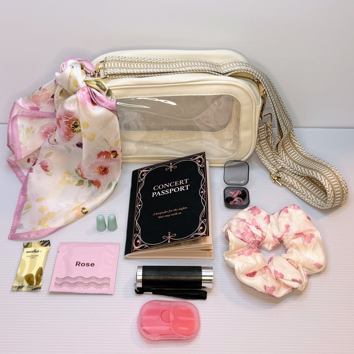 A curated flat-lay of a cream-colored cosmetic bag with a clear window and a woven strap, surrounded by travel accessories including a floral scarf, a black 'Concert Passport' booklet, a pink sample card, earplugs, a lip balm, a pink case, and two pink-and-white scrunchies on a light gray background.