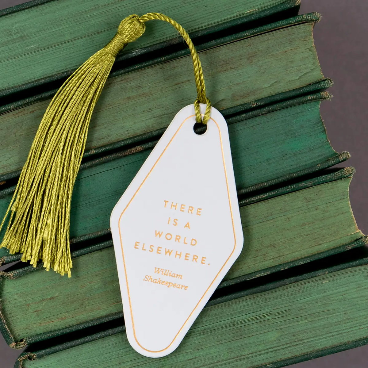White diamond-shaped vegan leather bookmark with gold foil typography reading There is a world elsewhere by William Shakespeare with gold silk tassel resting on vintage green books