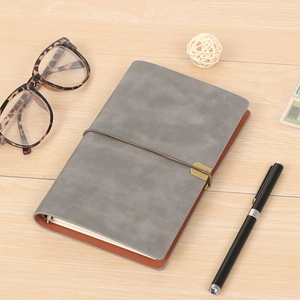 Gray vintage PU leather refillable journal with gold elastic strap closure, styled on white wood surface with tortoiseshell glasses and black pen