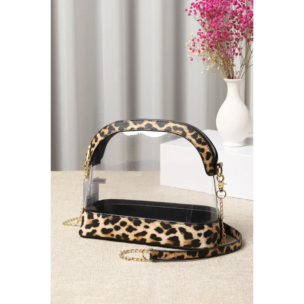 Large clear stadium bag with leopard print trim and gold chain strap styled on linen surface with pink flowers in background